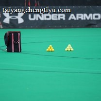 A sports field features a black and pink sports bag, two pyramids of yellow balls, and a portion of a person in dark athletic gear. The background displays a banner with the Under Armour logo.