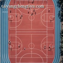 An aerial view of a large, vibrant sports complex featuring a red basketball court with multiple hoops and a surrounding blue running track. Several people are scattered across the area, engaged in various activities.