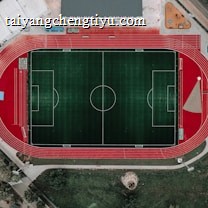 An aerial view of a large sports complex featuring a central green football field surrounded by a red running track. Adjacent to the track, there is a parking area, buildings, and pathways. Nearby, there are additional sports courts in different colors, including a blue swimming pool and orange basketball courts.