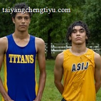 Two young athletes wearing sleeveless sports tops stand on a grassy field. The athlete on the left is wearing a blue top with the word 'TITANS' while the one on the right wears a yellow top with 'ASIJ'. The background features green trees and a white fence, with a bench visible on the right.