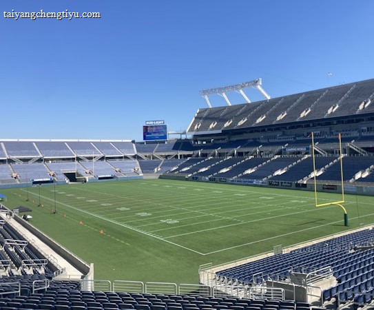 A large, empty sports stadium with blue seats surrounding a well-maintained green grass field. Bright sunlight creates clear shadows, and there are two goal posts on opposite ends of the field. A big electronic scoreboard displays some graphics, and the sky is clear and blue.